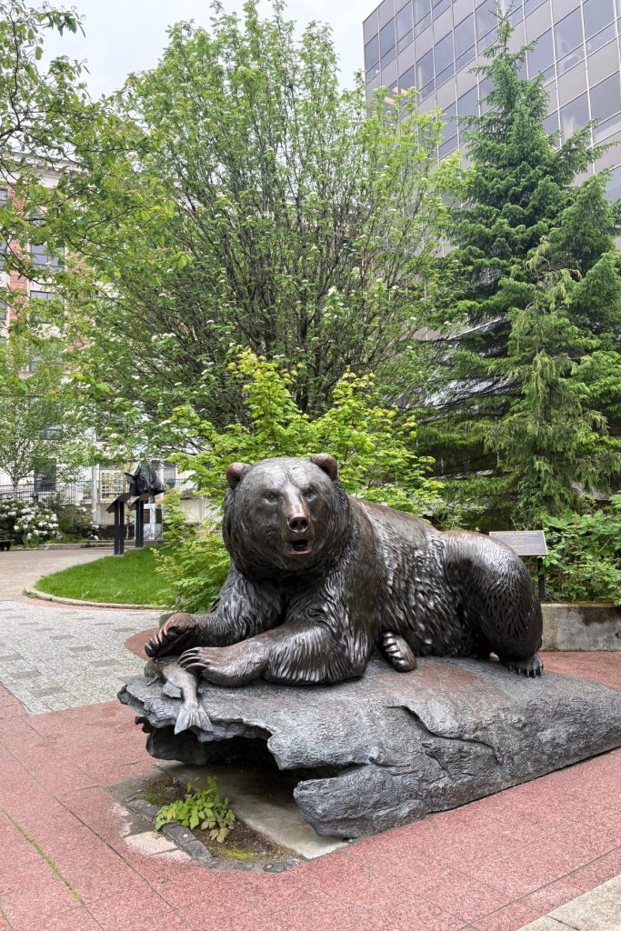 A bronze sculpture of a brown bear catching a salmon, titled Windfall Fisherman, resting on a rock outside a building in downtown Juneau.