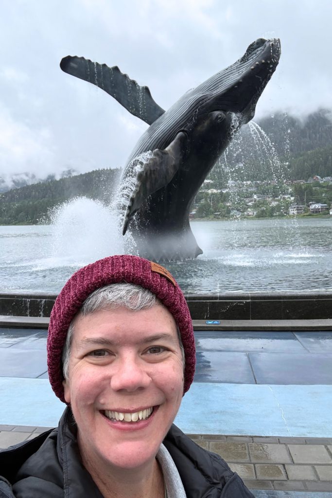 A woman smiling underneath Tahku, a bronze breaching humpback whale sculpture, as water mists around it at Overstreet Park in Juneau.