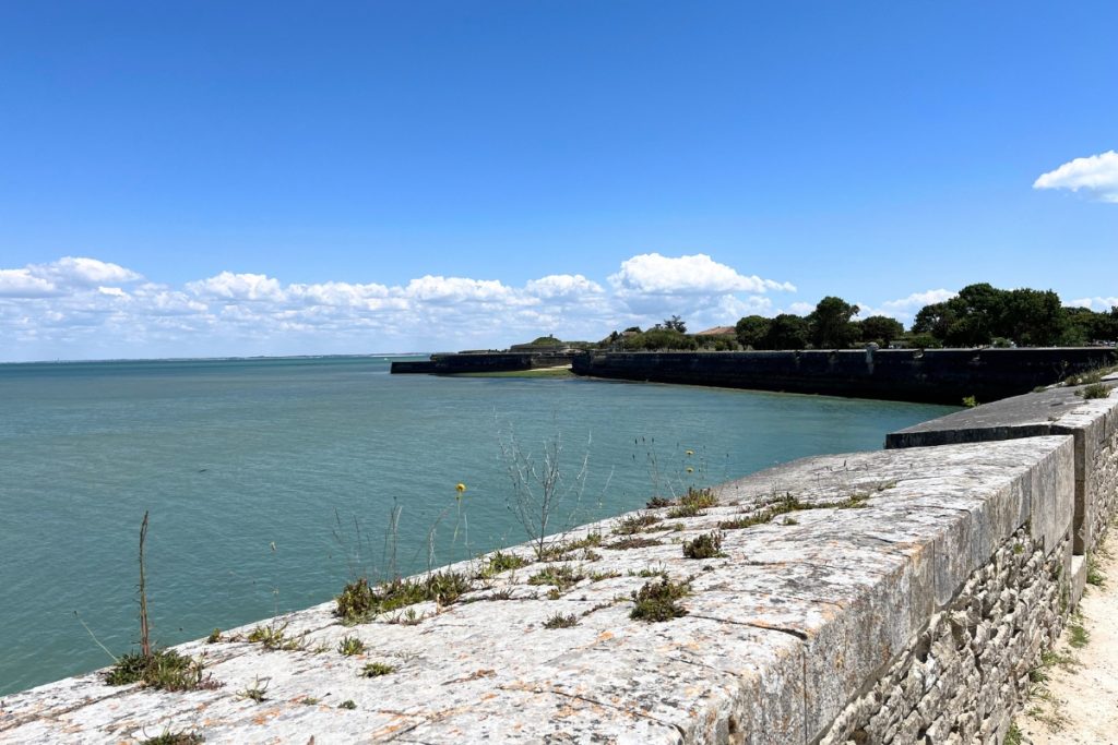 Stone rampart wall curving along the coastline in Saint-Martin-de-Ré, Île de Ré, France, with calm blue water and a distant fortified point.