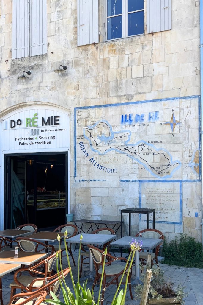 Hand painted map of Île de Ré, France, on a stone building facade above outdoor café tables, next to a bakery storefront sign reading "Do Ré Mie."