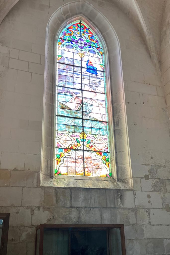 Tall Gothic stained glass window inside Église Saint-Martin, Île de Ré, France, depicting a sailing ship in blue and colorful floral borders.