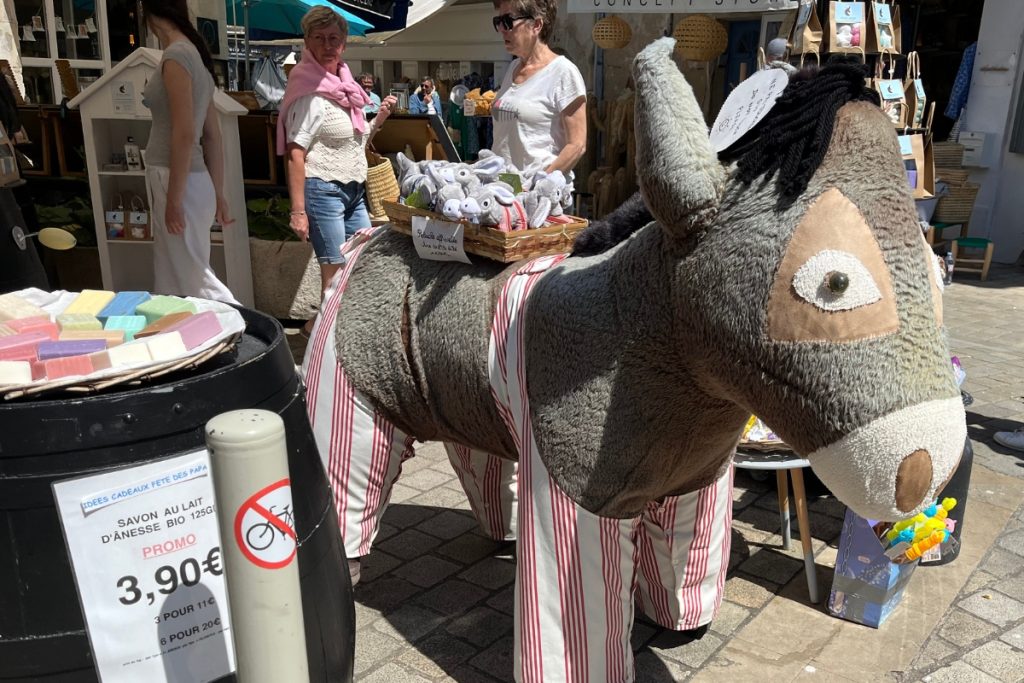 Life-size stuffed donkey mascot wearing red and white striped pants outside a shop selling donkey milk soap in Saint-Martin-de-Ré, Île de Ré, France, with shoppers browsing nearby.