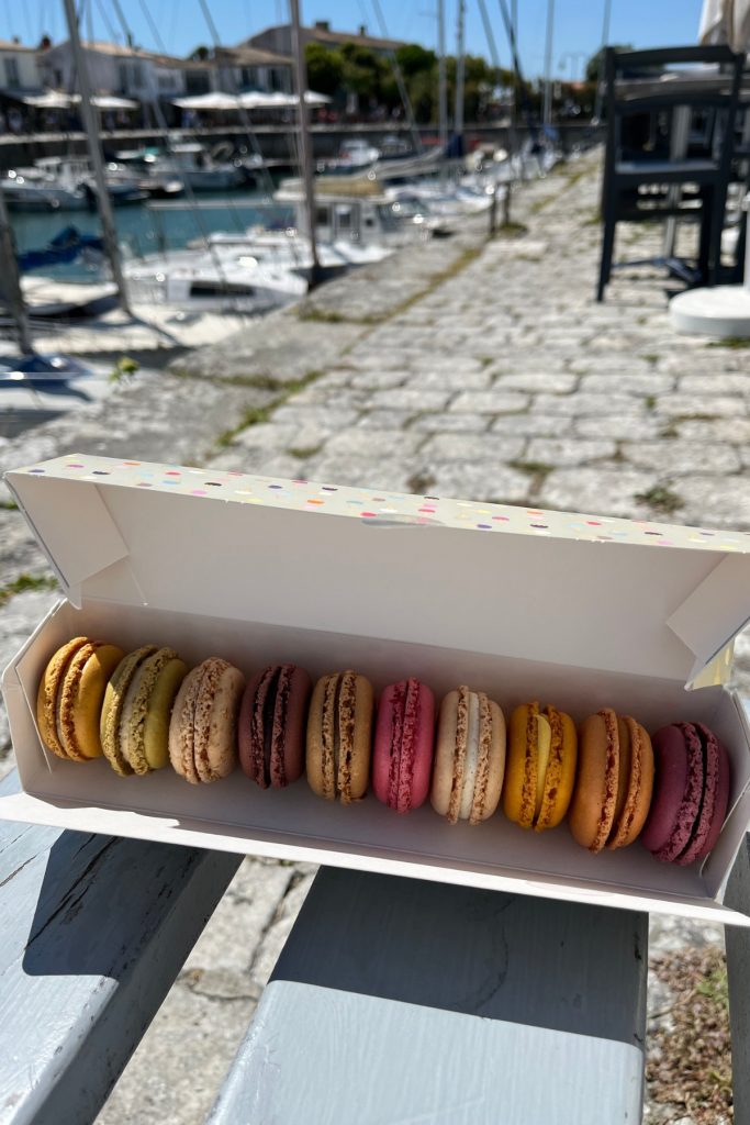 Open box of assorted French macarons in yellow, green, pink, and brown flavors on a wooden bench overlooking the boat harbor in Saint-Martin-de-Ré, Île de Ré, France.