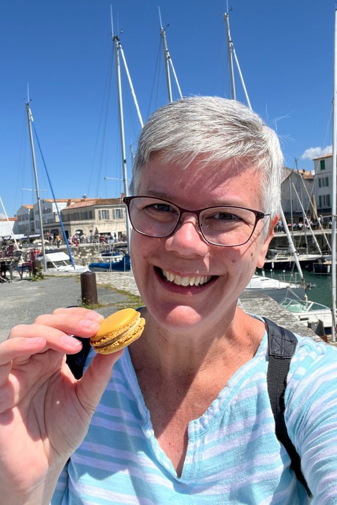Bethany smiling and holding a yellow macaron, with sailboat masts and the harbor of Saint-Martin-de-Ré, Île de Ré, France, in the background.