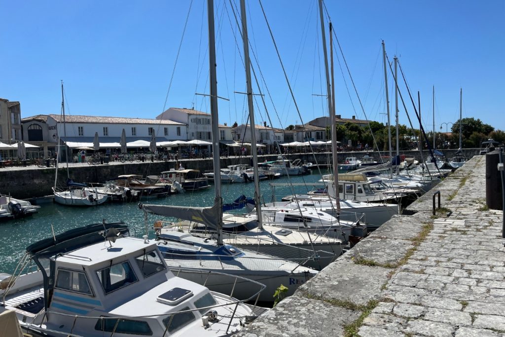 Sailboats and fishing boats moored in the stone harbor of Saint-Martin-de-Ré, Île de Ré, France, with white buildings and outdoor cafe umbrellas along the quay.