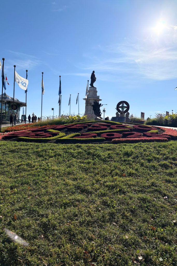 Landscaped flower bed with lettering, flags on poles, and a bronze monument in the background near Quebec City's Dufferin Terrace.