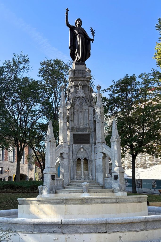 Gothic-style stone fountain topped by a bronze statue of a woman holding a cross, surrounded by trees in Quebec City.