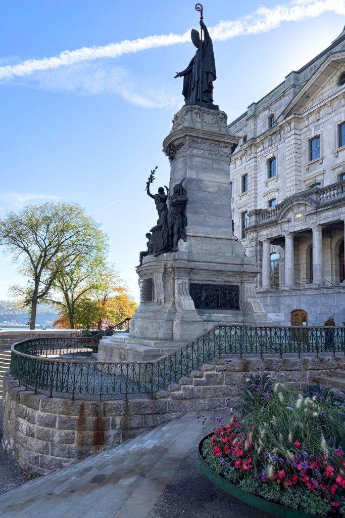 Bronze statue of a bishop atop a tall stone monument, beside a formal stone building with iron railings and flower beds.