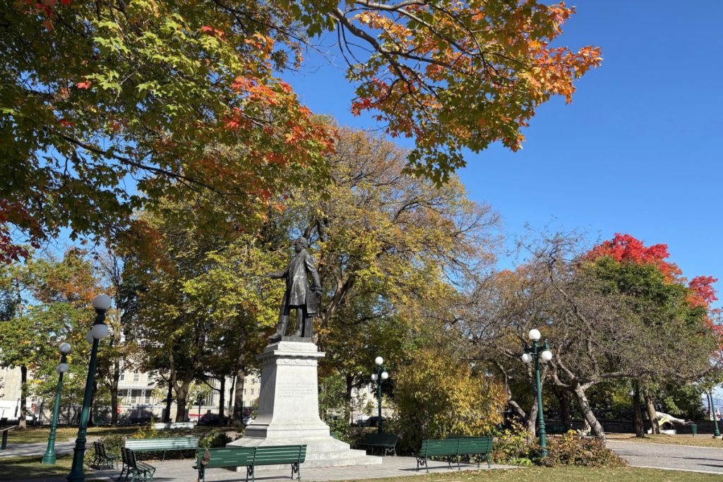 A bronze statue on a pedestal surrounded by green benches and trees in fall foliage at Parc Montmorency in Quebec City.
