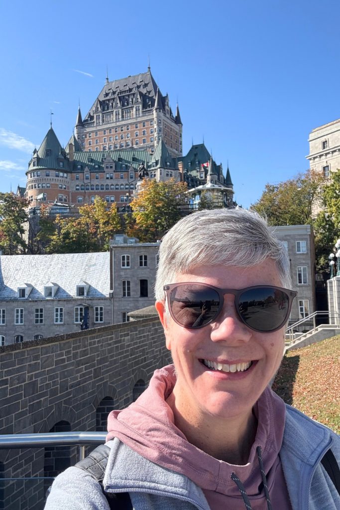 Selfie in sunglasses with the Château Frontenac hotel rising above stone buildings and fall foliage in the background.