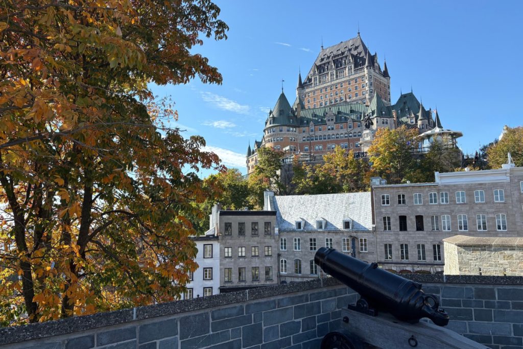 Château Frontenac hotel viewed from behind a black cannon at Parc Montmorency in Quebec City, with fall foliage in the foreground.