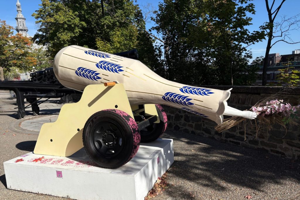 Cream-colored cannon art piece painted with blue wheat stalks, pink-patterned tires, and a hand holding flowers where the barrel's muzzle should be.