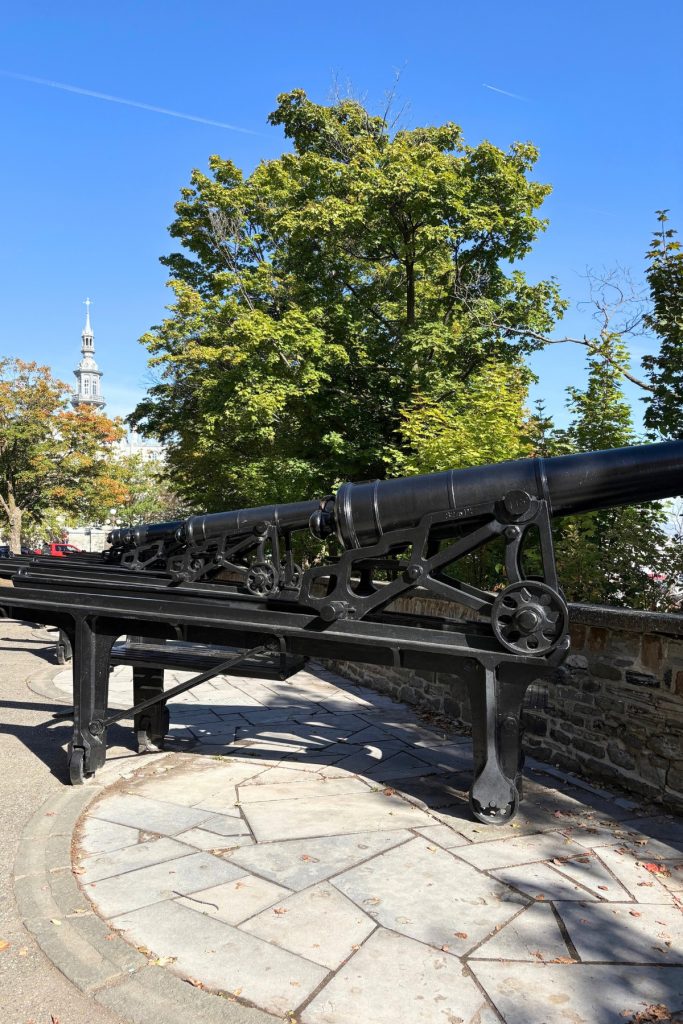 A row of black iron cannons on carriages pointed over a stone wall in Quebec City, with a church spire visible in the distance.