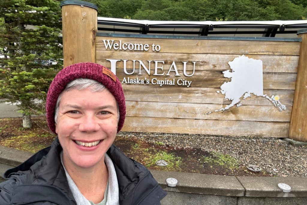 The same woman smiling in front of the Welcome to Juneau sign in 2026, wearing a different jacket.