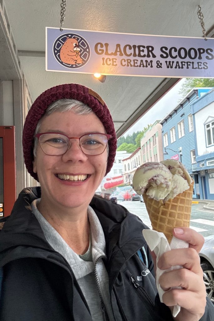 The same woman holding a waffle cone outside Glacier Scoops in downtown Juneau in 2026, wearing glasses and a different jacket.