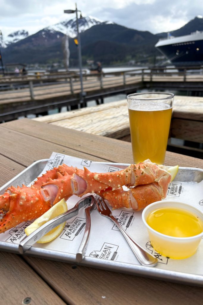 A tray of king crab legs with drawn butter and a glass of beer on a table at Alaskan Brewing Company Public House, with cruise ships visible in the background.