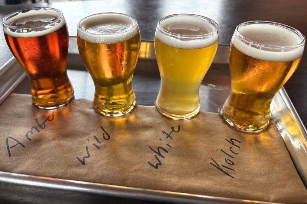 Four small glasses of beer in a flight, labeled Amber, Wild, White, and Kolsch, on a tray with a hand-lettered paper liner.