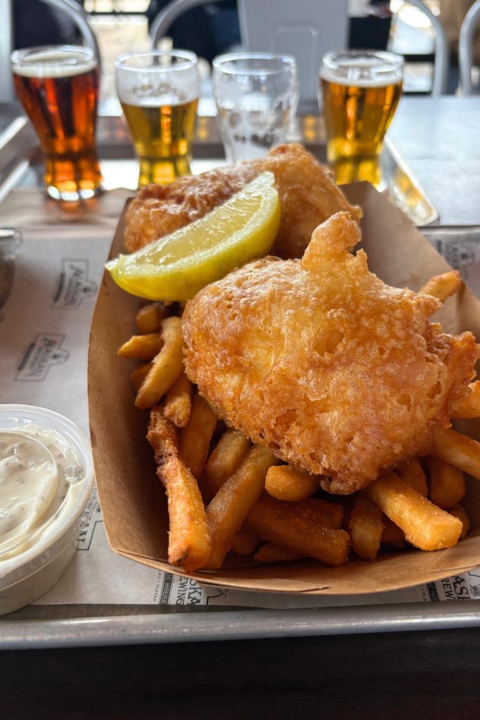 A basket of beer-battered fish and chips with a lemon wedge and tartar sauce, with a flight of four beers in the background.