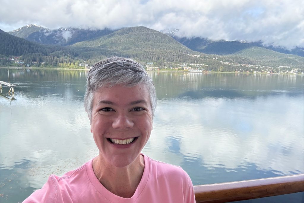 A woman smiling on a cruise ship deck with Juneau's harbor and mountains behind her, departing port in 2025.