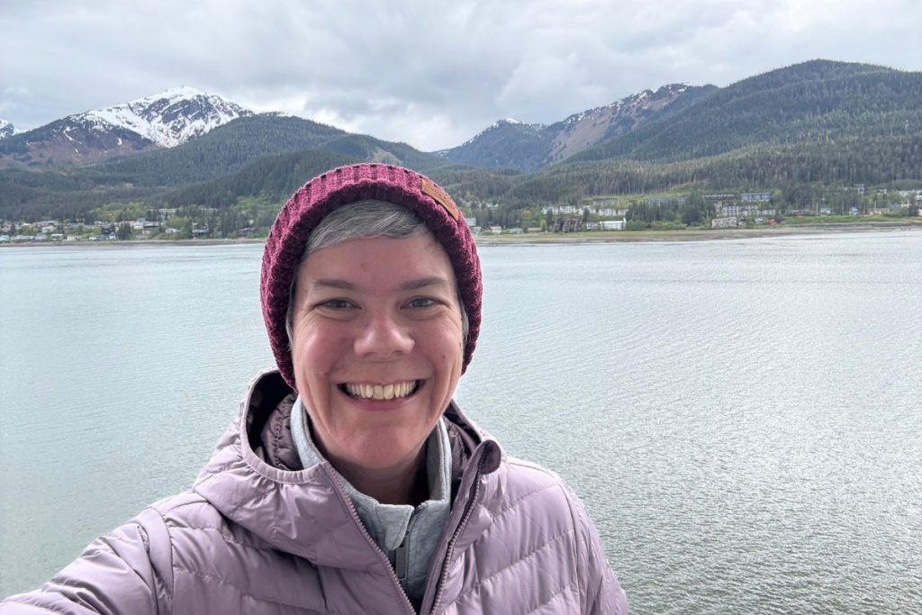 The same woman smiling on a cruise ship deck with Juneau's harbor and mountains behind her, departing port in 2026.