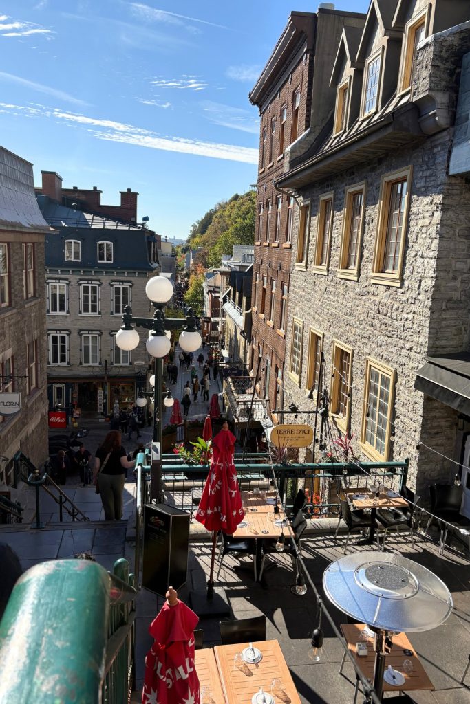 View down a Quebec City street lined with stone buildings, café tables, and a lamppost, seen from the top of the Breakneck Stairs.