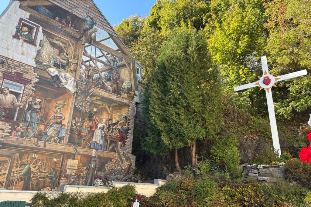 Trompe-l'oeil mural in Quebec City showing scenes of dockworkers and tradespeople, beside a white cross and garden greenery.