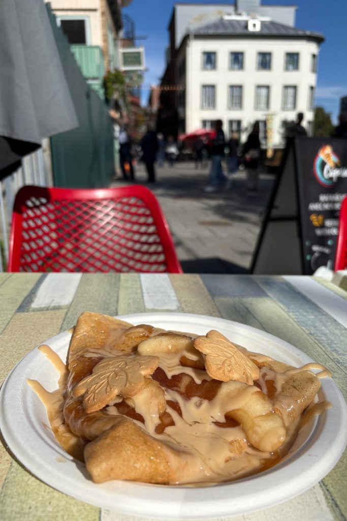 A crepe topped with apples, maple syrup, and maple leaf-shaped cookies on a paper plate, with a patio table and street scene in the background.