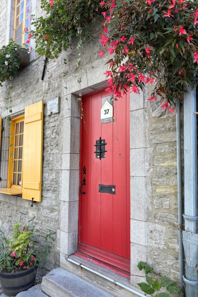 A red door numbered 37 on a stone building in Quebec City, framed by yellow shutters and hanging red begonias.