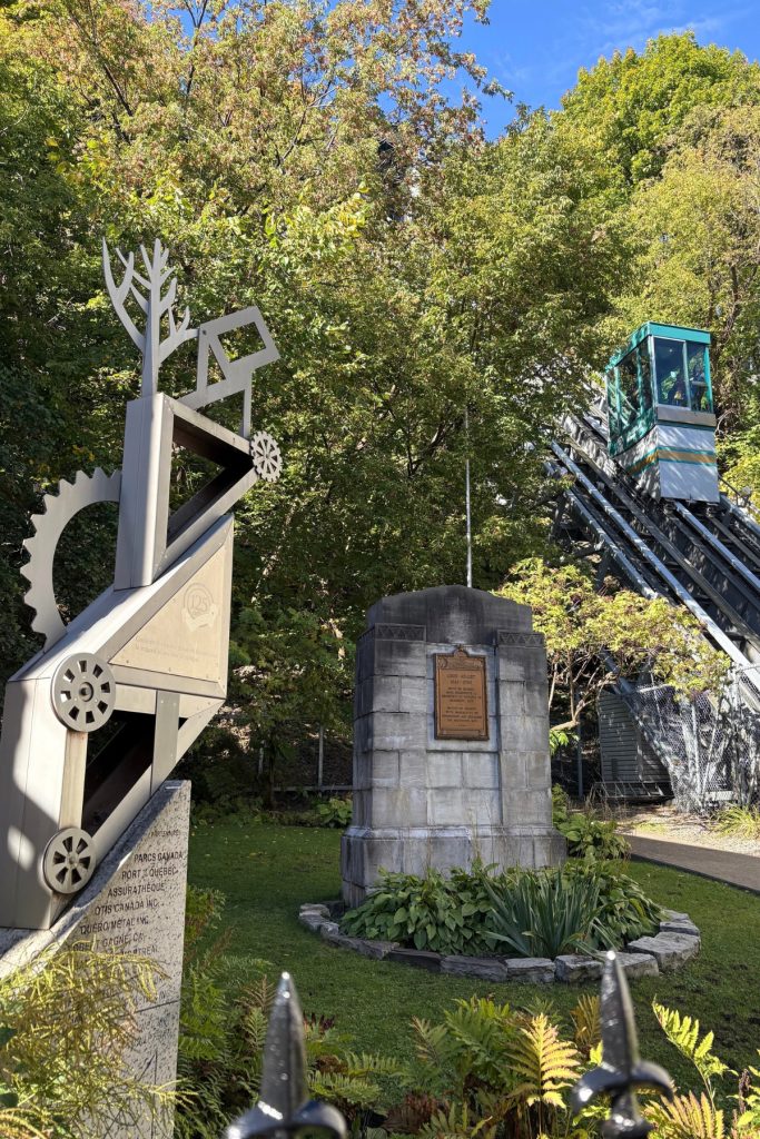 A stone marker with a bronze plaque and a metal sculpture beside the funicular tracks, with the funicular car descending through fall foliage in the background.
