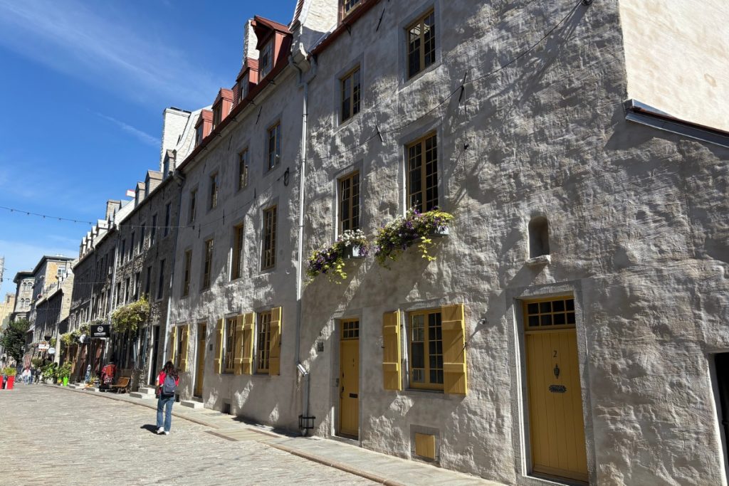 A row of whitewashed stone buildings with yellow shutters and doors along a cobblestone street in Quebec City, with a pedestrian walking past.