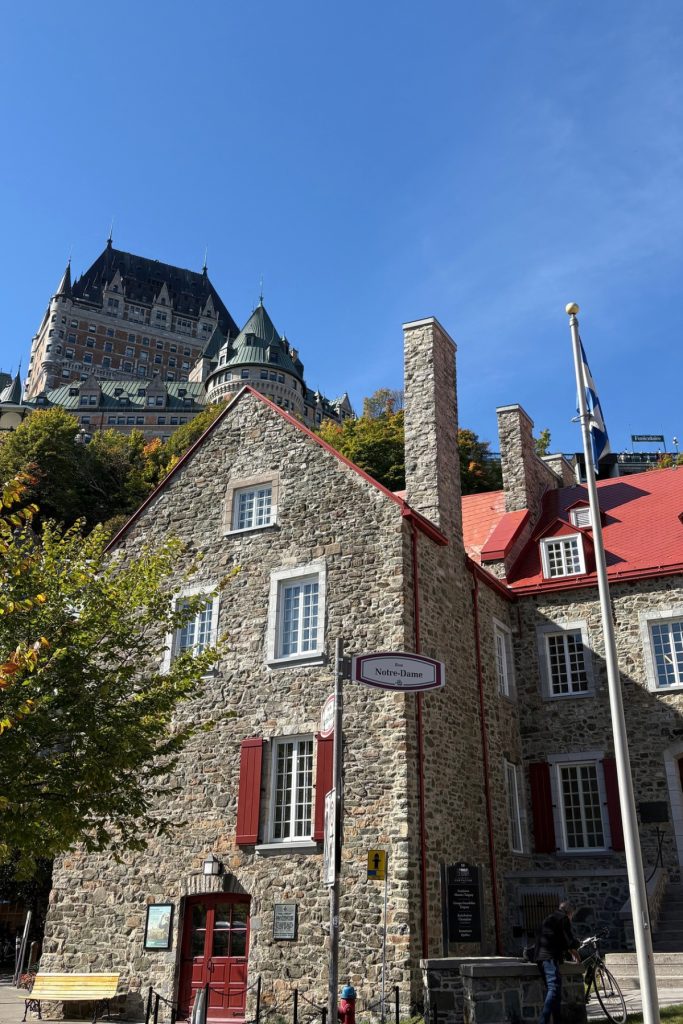 A stone building with red shutters, seen from below, with the Château Frontenac rising above it in the distance.