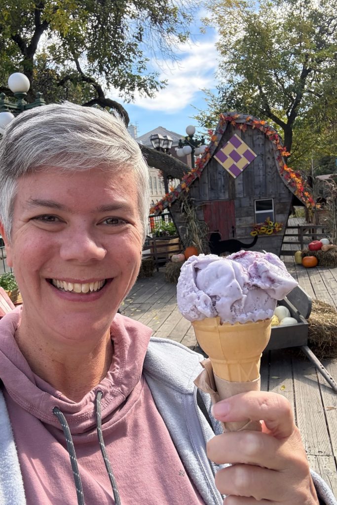 Selfie holding a blueberry ice cream cone in front of a rustic wooden fall display with pumpkins and a black cat silhouette.