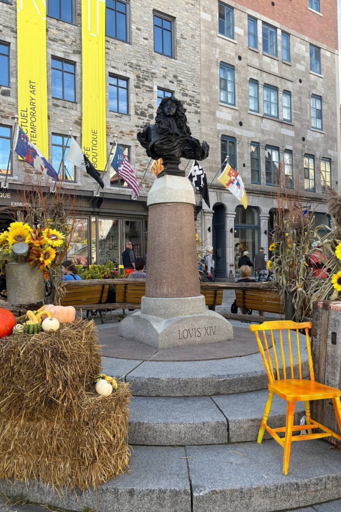 A bronze bust of Louis XIV on a stone pedestal, surrounded by hay bales, pumpkins, and an orange wooden chair.