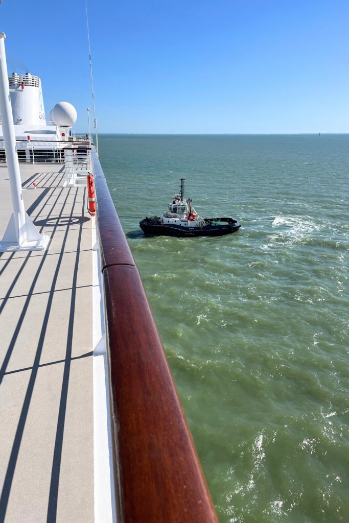 Tugboat assisting a cruise ship's departure from harbor on a windy day, viewed from the ship's deck railing, with green water and open sea in the background.