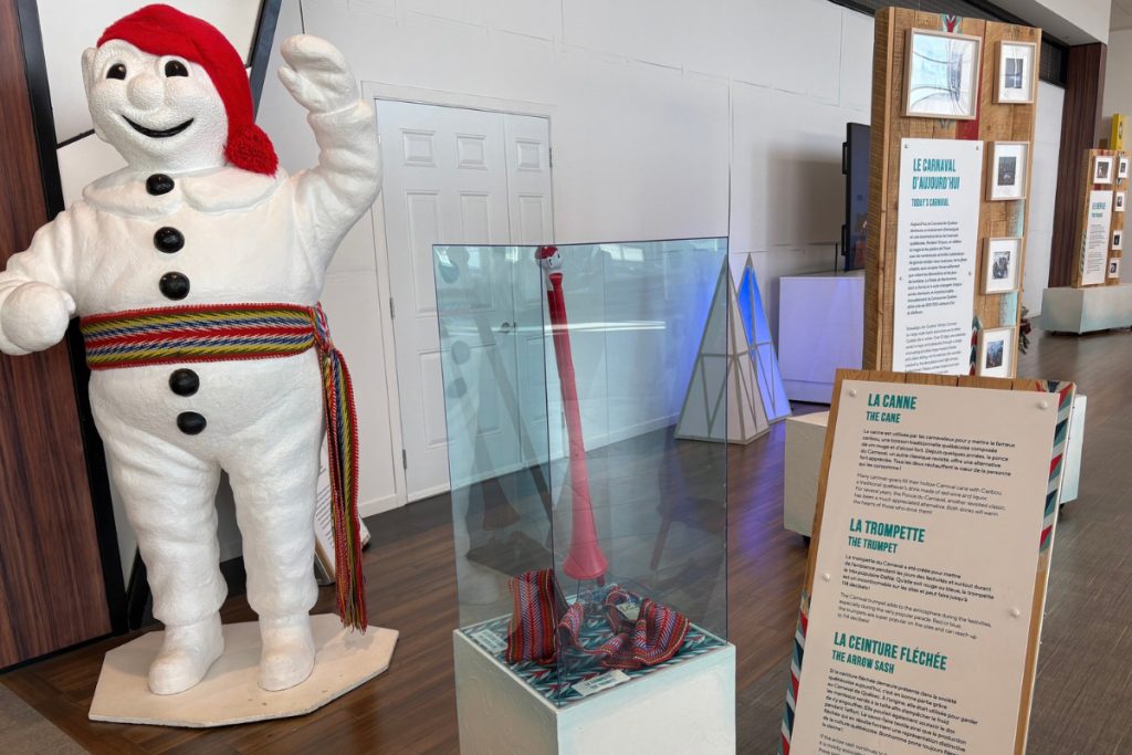 A large statue of Bonhomme Carnaval, a smiling snowman in a red cap and striped arrow sash, beside display cases at the Quebec City airport.