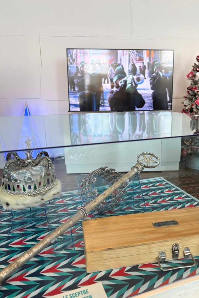 A jeweled ceremonial crown and scepter displayed in a glass case, with parade footage playing on a screen behind them.