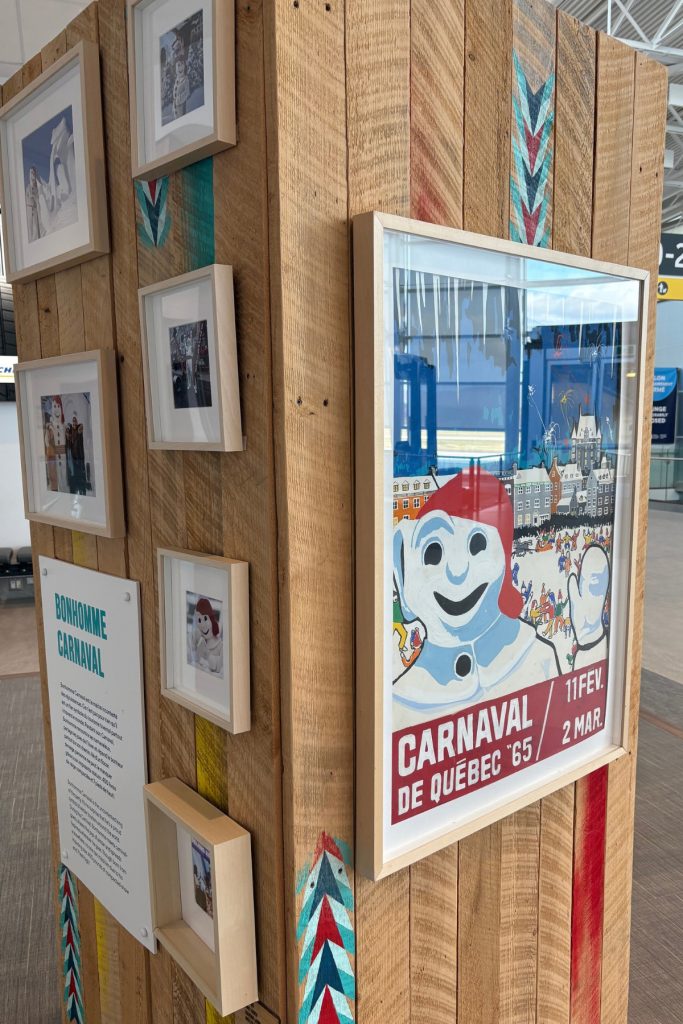 A framed 1965 Carnaval de Québec poster showing an illustrated Bonhomme Carnaval, displayed among photos on a wooden exhibit wall.