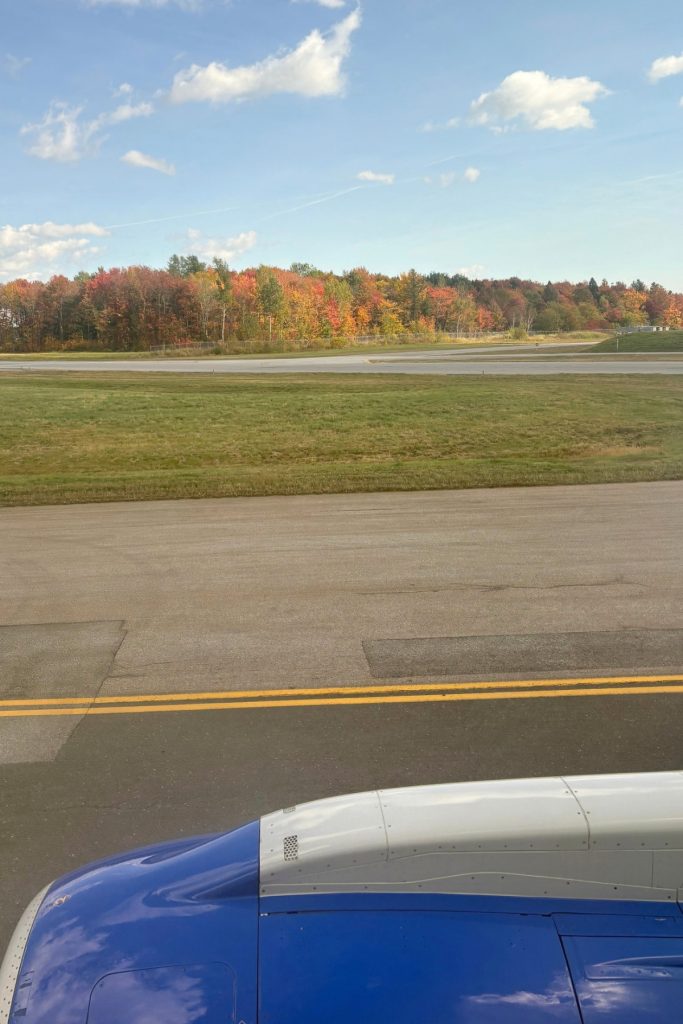 View of fall foliage along a runway from an airplane window, with part of the engine cowling visible in the foreground.