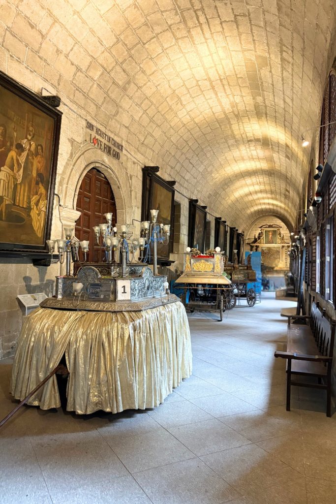 Vaulted stone corridor of the San Agustin Church cloister museum lined with gilded religious carrozas and portraits