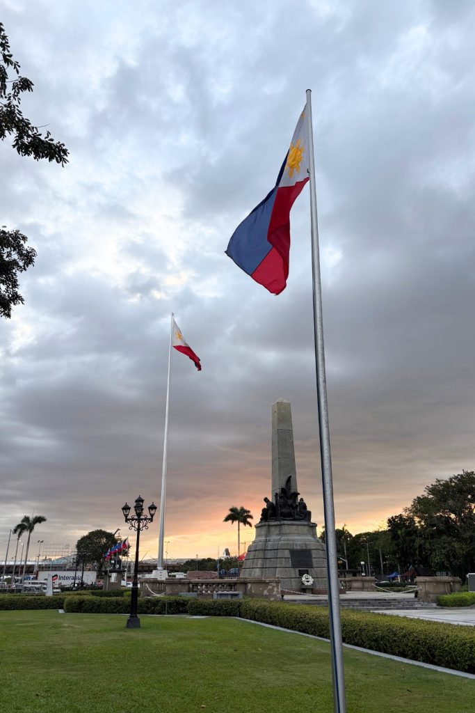 Philippine flags flying at sunset near the Rizal Monument obelisk at Rizal Park in Manila