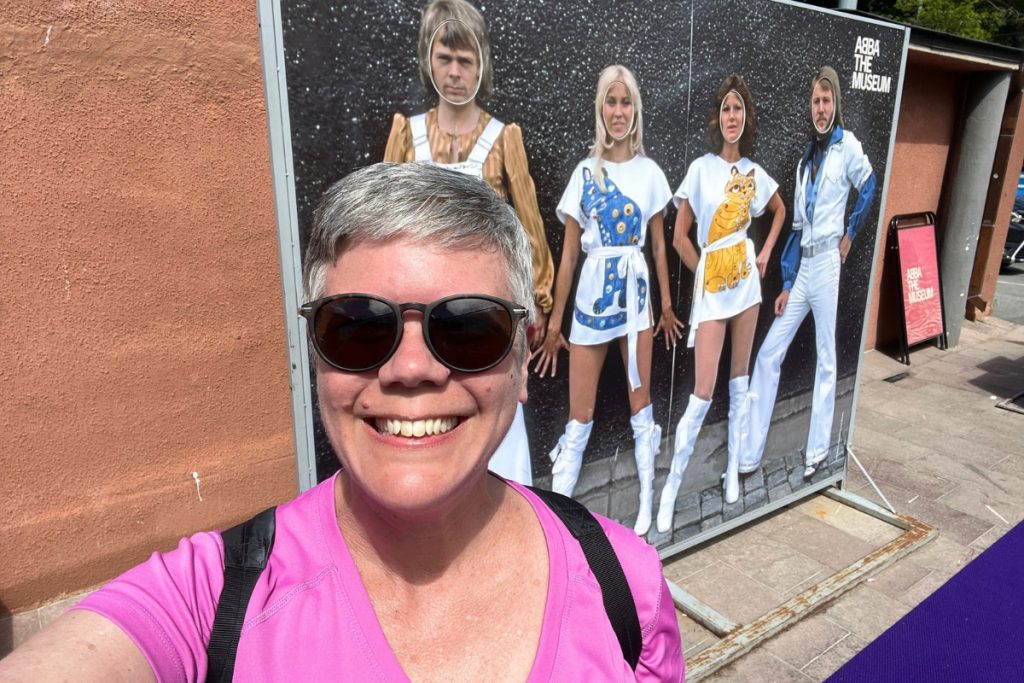 Woman with short gray hair and sunglasses taking a selfie in front of an ABBA The Museum display board in Stockholm.