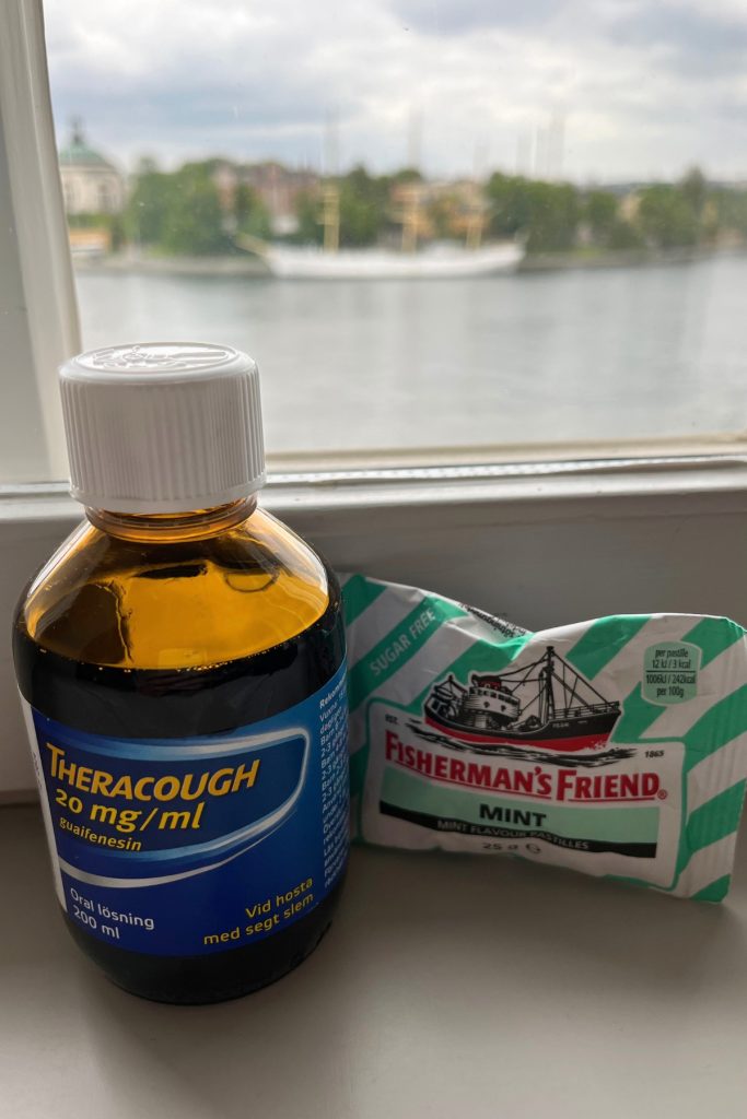 Bottle of Theracough cough syrup and a pack of Fisherman's Friend mint lozenges on a windowsill, with a harbor and a white sailing ship visible through the window.