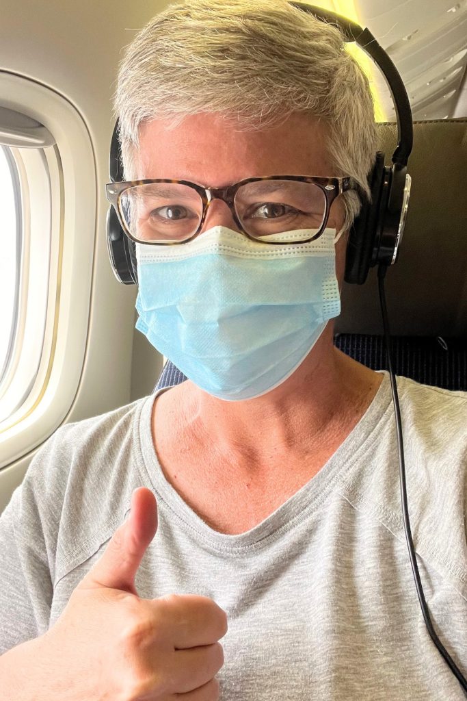 Woman in glasses wearing a blue surgical mask and over-ear headphones giving a thumbs up from an airplane window seat.