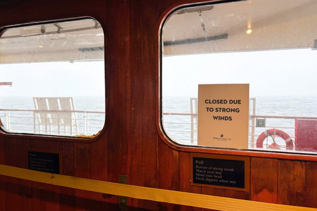 Sign taped inside a ship window reading Closed due to strong winds, with a life ring, an empty deck, and rough gray water beyond.